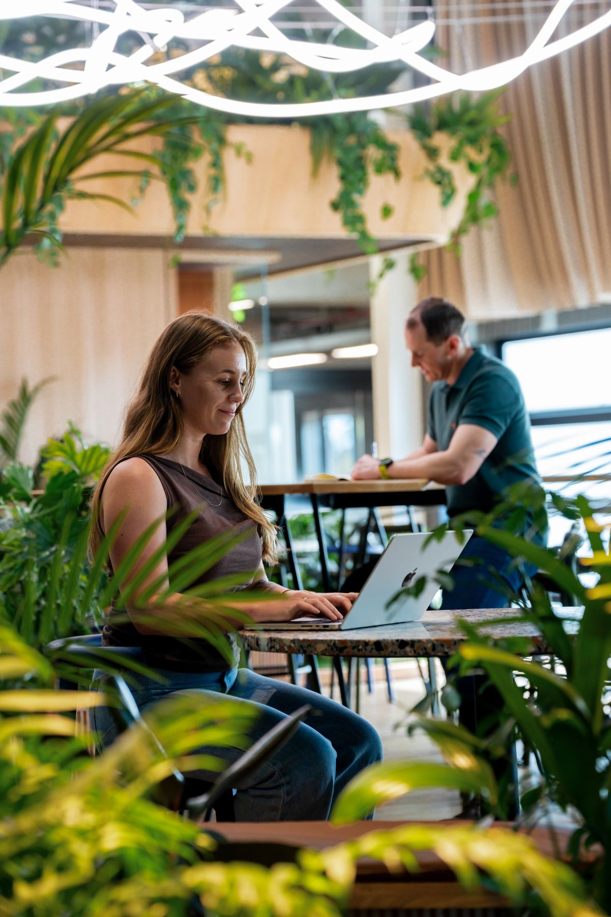 A woman working on a laptop surrounded by plants in a green, light-filled workspace